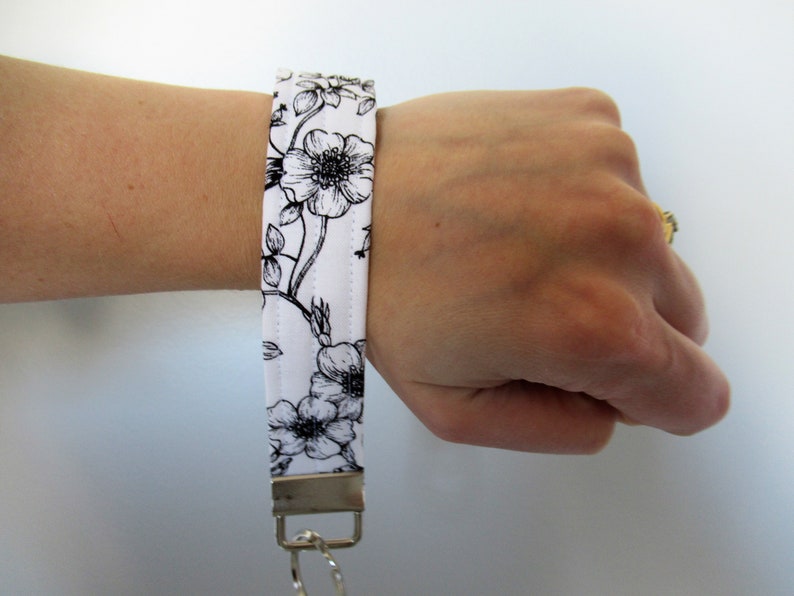 Op de afbeelding: Een witte sleutelhanger van stof met een zwarte bloemenprint en een zilveren sleutelring.
