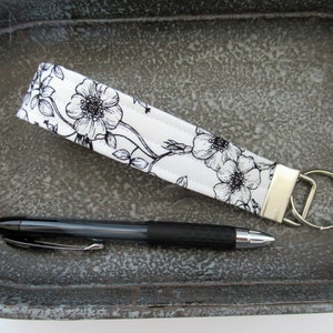 Op de afbeelding: Een witte stoffen sleutelhanger met een zwart bloemenpatroon en een zilveren sleutelring. De sleutelhanger ligt op een grijs, getextureerd oppervlak met een zwarte pen ernaast.