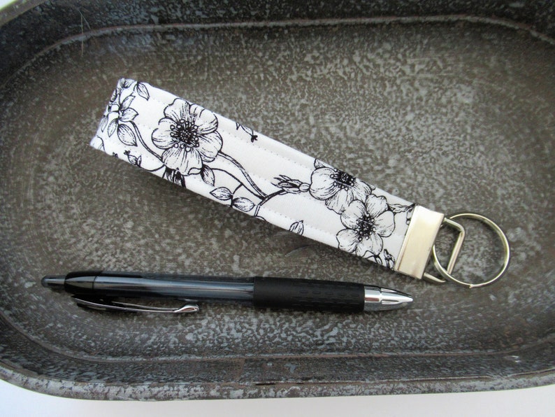 Op de afbeelding: Een witte stoffen sleutelhanger met een zwart bloemenpatroon en een zilveren sleutelring. De sleutelhanger ligt op een grijs, getextureerd oppervlak met een zwarte pen ernaast.