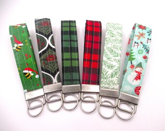 May include: Six fabric wrist keychains with silver-tone hardware. The patterns include green with bees in Santa hats, a black and olive design, green plaid, red plaid, white fern leaves, and a snowman with a Christmas tree.