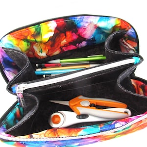 May include: Open, multi-compartment pencil case with a vibrant, abstract watercolor pattern in shades of blue, pink, yellow, and green. The interior is black, and it contains pens, scissors, and a rotary cutter. The case has a white zipper.