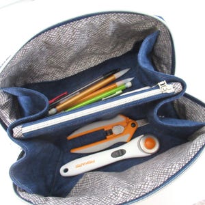 May include: A navy blue fabric organizer bag with a zippered central compartment and open side sections. The interior is lined with a grey and white crosshatch pattern. It contains several pens, orange and grey snips, and a white rotary cutter.