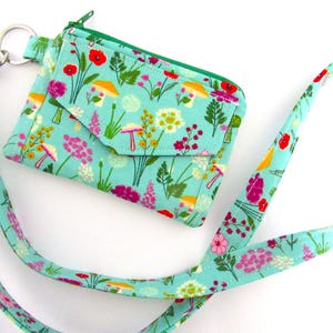 May include: A teal fabric wallet with a matching lanyard, both adorned with a floral and mushroom pattern. The wallet has a flap closure and a green zipper. The lanyard is connected to the wallet with a silver-colored clasp.
