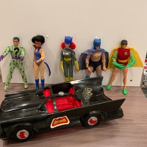 May include: A collection of five action figures including The Riddler, Catwoman, Batgirl, Batman, and Robin, standing behind a black toy car with a red interior and a Batman logo on the side. The figures feature fabric costumes and plastic accessories.