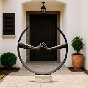 May include: A bronze sculpture depicting two hands clasped within a dark metal circle. The sculpture is mounted on a rectangular stone base. The artwork is positioned in front of a dark wooden door and a white exterior wall.
