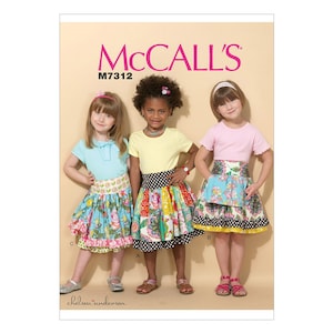 May include: McCall's sewing pattern M7312 for girls' dresses. The pattern includes three different styles: a blue dress with a floral print, a yellow dress with a patchwork print, and a pink dress with a floral print. Each dress has a ruffled skirt and a contrasting waistband.