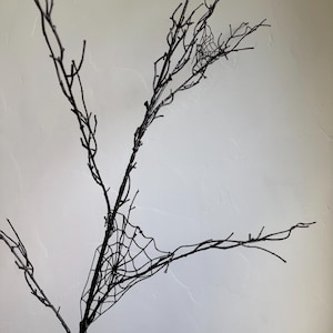 May include: A black decorative branch featuring multiple thin, bare twigs extending outward. Attached to the branch are two distinct, intricate black spider web designs, one positioned lower on the main stem and a smaller one near the top.