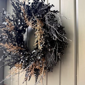 May include: A circular wreath composed of black foliage, dark leaves, and dried brown twigs. The arrangement features thin, bare white branches extending outward, creating a textured, layered appearance against a solid, paneled door surface.