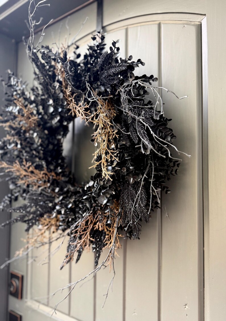 May include: A circular wreath composed of black foliage, dark leaves, and dried brown twigs. The arrangement features thin, bare white branches extending outward, creating a textured, layered appearance against a solid, paneled door surface.
