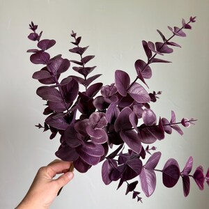 May include: A cluster of artificial eucalyptus stems featuring deep purple, rounded leaves arranged along thin, branching stalks. The foliage displays a matte texture with visible vein patterns on each leaf, creating a dense, layered appearance.