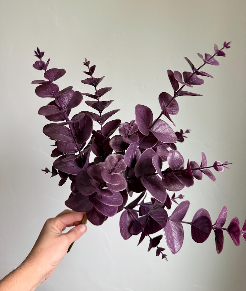 May include: A cluster of artificial eucalyptus stems featuring deep purple, rounded leaves arranged along thin, branching stalks. The foliage displays a matte texture with visible vein patterns on each leaf, creating a dense, layered appearance.