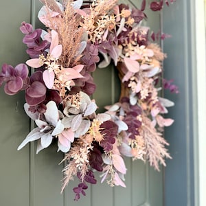 May include: A circular wreath featuring a mix of dried and faux botanical elements in shades of deep plum, dusty rose, and muted sage green. The arrangement includes eucalyptus leaves, feathery pampas grass, and small floral sprigs.
