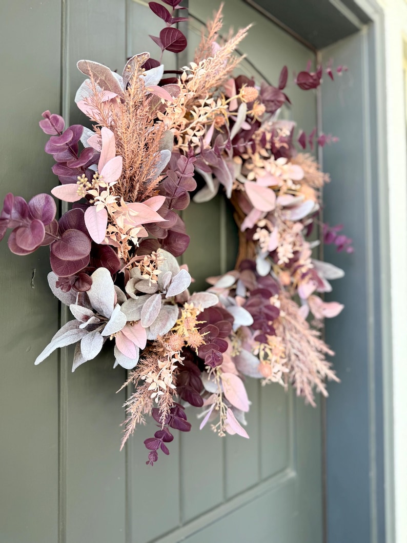 May include: A circular wreath featuring a mix of dried and faux botanical elements in shades of deep plum, dusty rose, and muted sage green. The arrangement includes eucalyptus leaves, feathery pampas grass, and small floral sprigs.