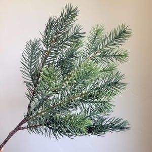 May include: A single artificial pine branch featuring multiple stems with needle-like foliage in shades of muted sage green and deep forest green. The main stem is textured and brown, branching out into several smaller sprigs of evergreen needles.