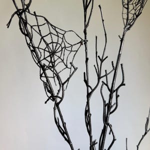 May include: A set of bare, black-painted branches featuring a delicate, glittery spider web design attached between the twigs. The branches are thin and spindly, with a dark, matte finish that contrasts with the shimmering, intricate web pattern.