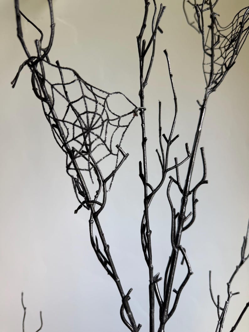 May include: A set of bare, black-painted branches featuring a delicate, glittery spider web design attached between the twigs. The branches are thin and spindly, with a dark, matte finish that contrasts with the shimmering, intricate web pattern.