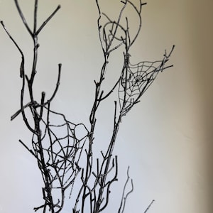 May include: A collection of bare, black-painted branches featuring two delicate, intricate spider web designs attached to the twigs. The branches are thin, multi-directional, and have a smooth, dark finish throughout the entire structure.