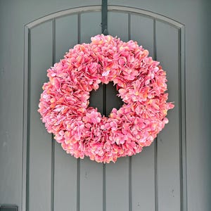 May include: A vibrant pink hydrangea wreath hangs on a gray door. The wreath is circular, densely packed with layered petals, and features a mix of pink and peach hues. A black metal hanger supports the wreath.