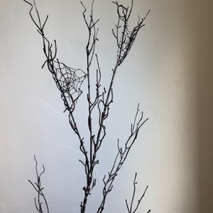 May include: A tall, slender, black decorative branch featuring multiple thin, bare twigs extending upward. Two delicate, white, intricate spider web patterns are attached to the upper sections of the branches, creating a stark contrast against the dark wood.
