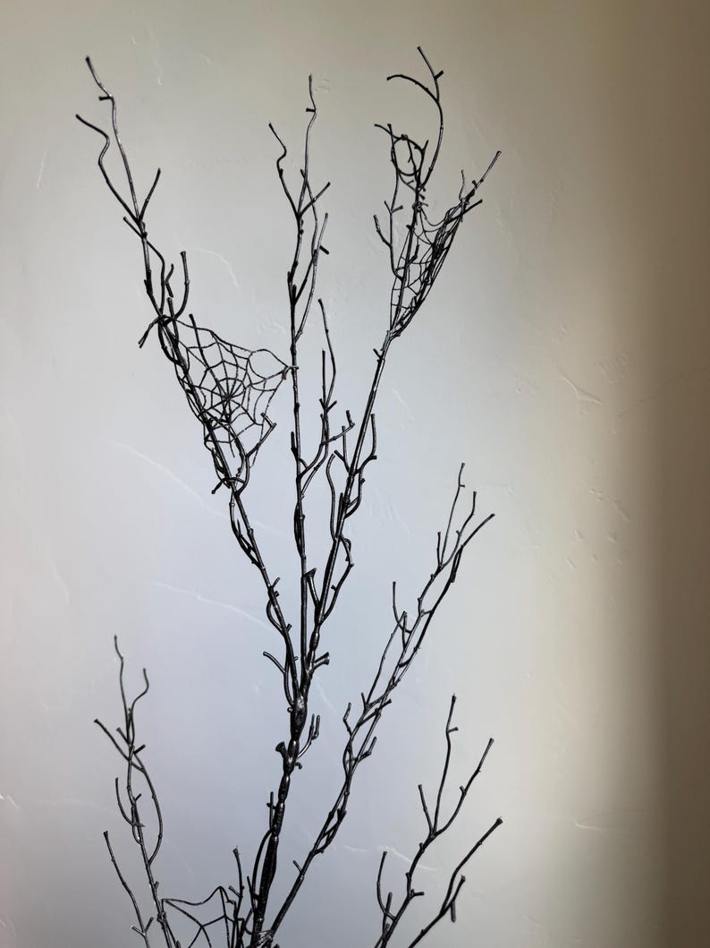 May include: A tall, slender, black decorative branch featuring multiple thin, bare twigs extending upward. Two delicate, white, intricate spider web patterns are attached to the upper sections of the branches, creating a stark contrast against the dark wood.