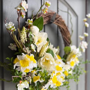 May include: A spring wreath with a natural grapevine base, adorned with white and yellow daffodils, white blossoms, and green foliage. The wreath is hanging on a gray door, creating a welcoming seasonal decoration.