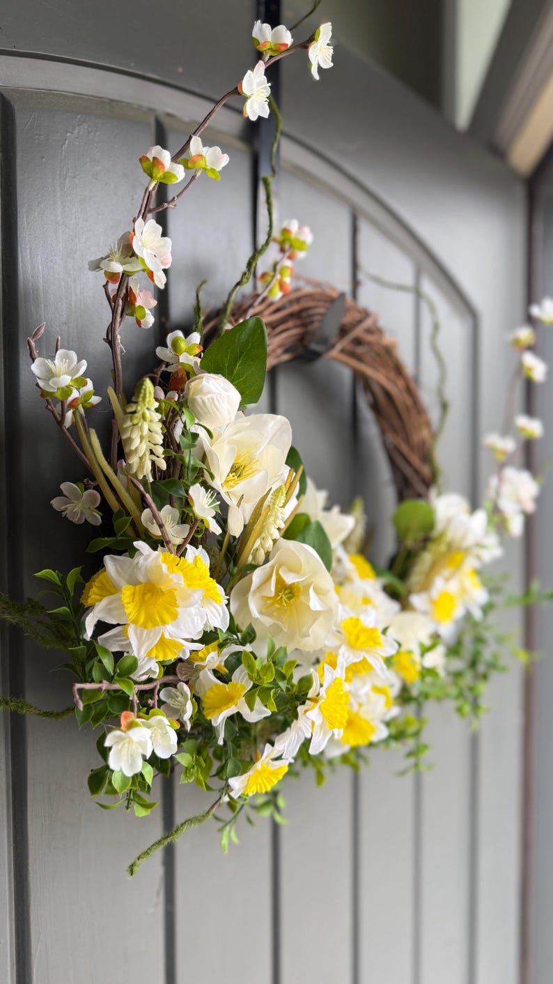 May include: A spring wreath with a natural grapevine base, adorned with white and yellow daffodils, white blossoms, and green foliage. The wreath is hanging on a gray door, creating a welcoming seasonal decoration.