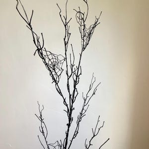 May include: A tall, slender decorative branch painted in matte black, featuring multiple thin, twig-like extensions. Several delicate, white, web-like structures are attached to the branches, creating a stark contrast against the dark wood.
