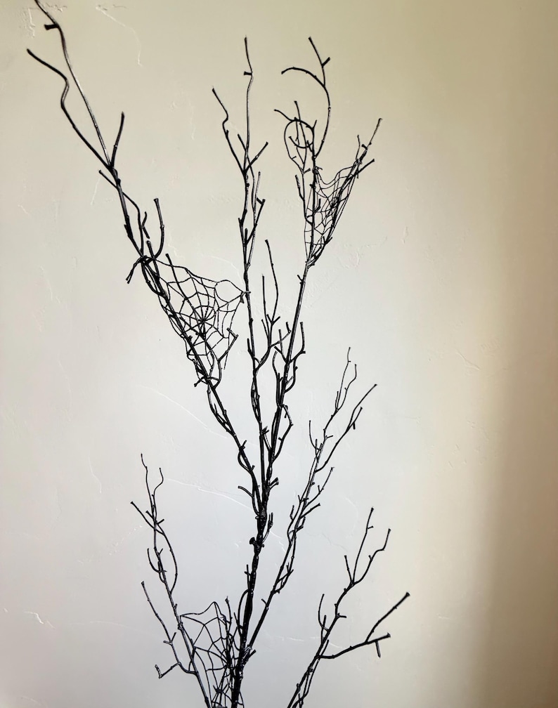 May include: A tall, slender decorative branch painted in matte black, featuring multiple thin, twig-like extensions. Several delicate, white, web-like structures are attached to the branches, creating a stark contrast against the dark wood.