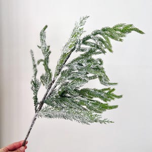 May include: A single artificial evergreen branch featuring vibrant green needle-like foliage coated with a white, frost-like texture. The stem is dark brown and slender, supporting multiple smaller, branching sprigs that extend outward in a natural, asymmetrical arrangement.