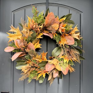 May include: A circular wreath featuring a mix of artificial autumn foliage, including yellow and orange maple leaves, green magnolia leaves, and brown pinecones. The arrangement is accented with sprigs of evergreen needles and dried cedar branches.