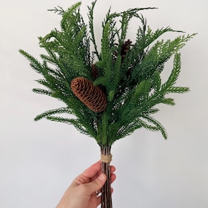 May include: A decorative bouquet of artificial green cedar branches and pine needles featuring two brown pinecones. The stems are gathered together and secured with a piece of natural twine, creating a bundle of evergreen foliage.