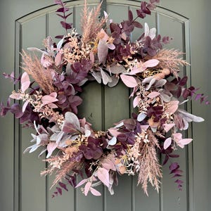 May include: A circular wreath featuring a mix of deep plum, dusty rose, and soft grey foliage. The arrangement includes eucalyptus leaves, feathery pampas grass plumes, and small dried floral accents, creating a layered, textured botanical display.