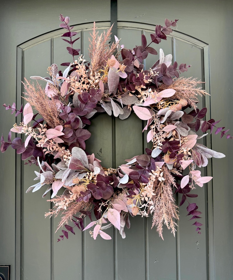May include: A circular wreath featuring a mix of deep plum, dusty rose, and soft grey foliage. The arrangement includes eucalyptus leaves, feathery pampas grass plumes, and small dried floral accents, creating a layered, textured botanical display.