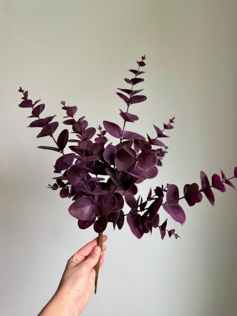 May include: A decorative artificial eucalyptus branch featuring multiple stems with rounded, deep purple leaves. The foliage is arranged in a branching pattern, with smaller leaves near the tips and larger, more mature leaves toward the base.