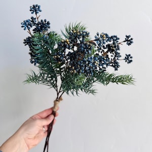 May include: A decorative floral spray featuring clusters of dark blue berries with subtle gold accents, interspersed with green pine-like needles. The stems are gathered together and secured with a light brown twine wrapping at the base.