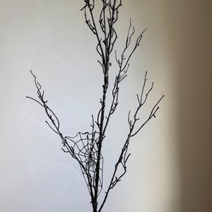 May include: A decorative black branch featuring multiple thin, bare twigs extending from a central stem. Two distinct, intricate spider web designs are attached to the branches, one positioned near the top and another on a lower side branch.