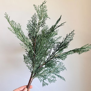 May include: A faux cedar branch featuring multiple stems with dense, needle-like foliage in a muted sage green color. The branch has a brown, textured main stem that branches out into several smaller, delicate sprigs of evergreen-style greenery.
