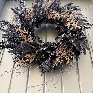 May include: A circular wreath composed of dark black foliage and interspersed with light brown, dried twig-like branches. The arrangement features a dense collection of leaves and thin, spindly stems that extend outward from the central ring structure.