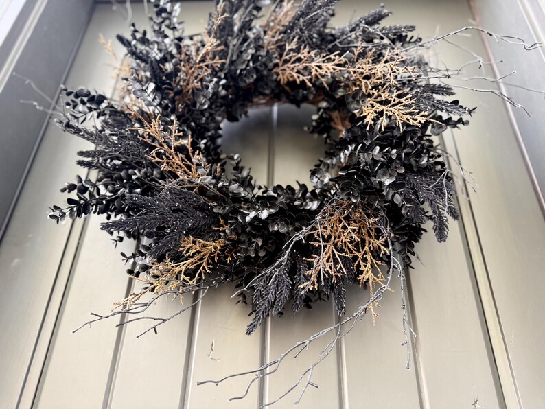 May include: A circular wreath composed of dark black foliage and interspersed with light brown, dried twig-like branches. The arrangement features a dense collection of leaves and thin, spindly stems that extend outward from the central ring structure.