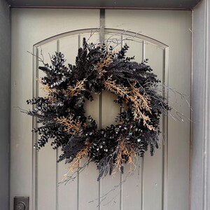 May include: A circular wreath composed of dark black foliage, including eucalyptus leaves and thin twigs, accented with contrasting light brown, dried natural branches. The arrangement features a dense, layered texture with protruding stems.