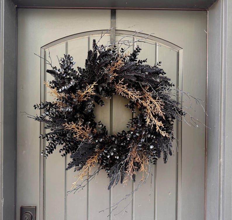 May include: A circular wreath composed of dark black foliage, including eucalyptus leaves and thin twigs, accented with contrasting light brown, dried natural branches. The arrangement features a dense, layered texture with protruding stems.