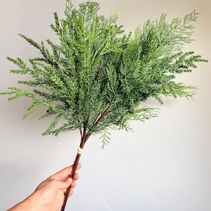 May include: A bundle of artificial cedar branches featuring vibrant green, needle-like foliage and textured brown stems. The branches are gathered together and secured with light-colored twine at two points along the lower stems.