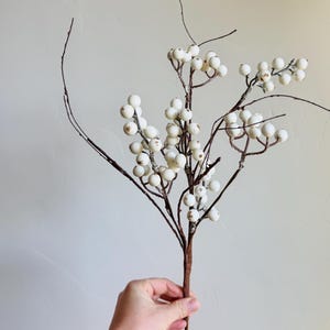 May include: A decorative artificial stem featuring clusters of white, round berries. The berries are attached to thin, brown branches that extend from a central stem. The stem is held against a neutral background, showcasing the naturalistic design.