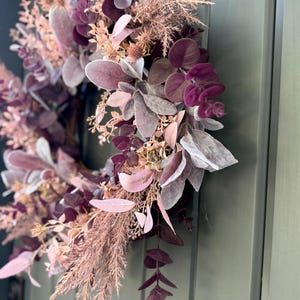 May include: A circular wreath featuring a dense arrangement of faux foliage in shades of deep plum, dusty rose, and muted mauve. The design incorporates rounded eucalyptus leaves, feathery grasses, and soft, velvety lamb's ear foliage.