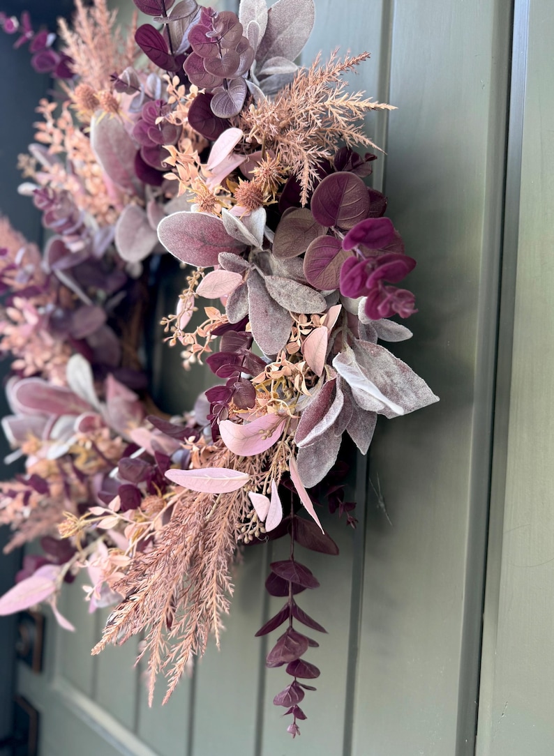May include: A circular wreath featuring a dense arrangement of faux foliage in shades of deep plum, dusty rose, and muted mauve. The design incorporates rounded eucalyptus leaves, feathery grasses, and soft, velvety lamb's ear foliage.