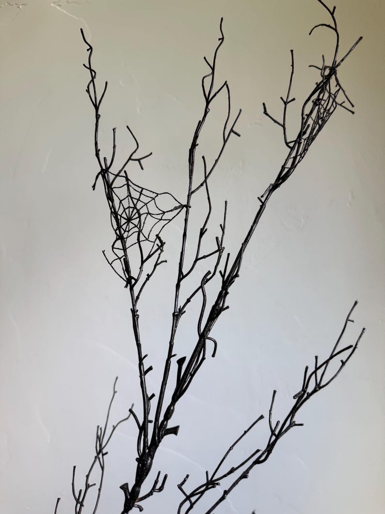 May include: A set of bare, black-painted branches featuring thin, intricate, black spider web designs attached to the twigs. The branches are slender and multi-pronged, extending upward with a dark, matte finish throughout the structure.