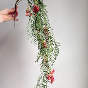 May include: A decorative, artificial hanging plant with long, slender green leaves and clusters of small, red berries. The plant is held by a hand against a neutral background. The plant's curved shape adds a graceful touch.