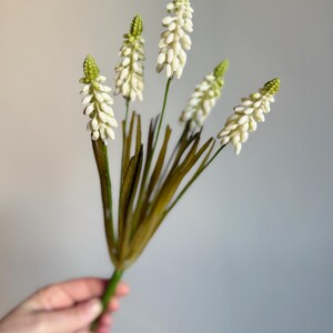 May include: A bundle of artificial flowers with white, clustered blooms and green tips. The stems are green and brown, with long, thin leaves. The flowers are held against a plain gray background.