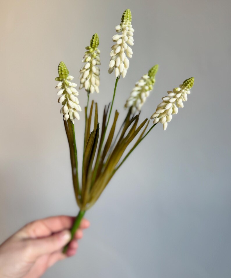 May include: A bundle of artificial flowers with white, clustered blooms and green tips. The stems are green and brown, with long, thin leaves. The flowers are held against a plain gray background.