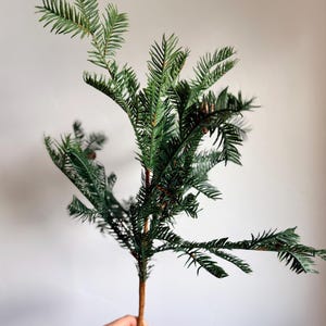 May include: A single artificial evergreen branch featuring dark green, needle-like foliage arranged on a central brown stem. The branch has multiple smaller offshoots extending outward, creating a realistic, layered appearance of pine needles.